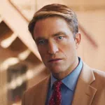 Man in a tan suit and red polka dot tie staring directly at the camera with a blurred warm background.