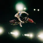 Motocross rider performing a stunt mid-air against bright stadium lights in a dark arena.