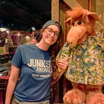 Smiling woman wearing glasses and a cap poses with ALF character doll in a tropical shirt inside a dimly lit venue.