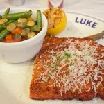 Breaded meat cutlet topped with grated cheese and herbs served with mixed vegetables and grilled lemon on a white plate.