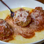 Three meatballs topped with grated cheese served in marinara sauce over creamy polenta in a white bowl.