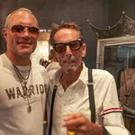 Two men wearing sunglasses and necklaces posing indoors at a jewelry store with display cases behind them