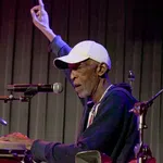 Elderly male musician playing congas and singing into a microphone with one hand raised on stage.