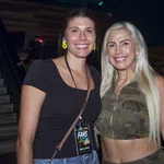 Two smiling women posing indoors, one in black top and the other in camouflage crop top with long blonde hair.