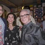 Three adults smiling together indoors, dressed in semi-formal attire at a social gathering with framed art on walls.