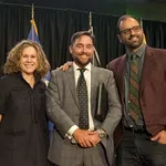 Three professionally dressed people smiling with flags in the background at an indoor event.