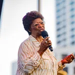 Elderly woman in striped blouse speaking into microphone at outdoor event with city building background