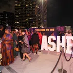 People dressed in elegant attire gather at a nighttime outdoor event with large illuminated ASHE letters and city buildings in the background