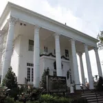 White Greek Revival mansion with tall Corinthian columns and manicured garden in front under a clear sky.