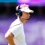 Football coach wearing LSU visor and headset focused during a game with blurred purple stadium background