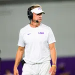 LSU football coach in white LSU gear and visor wears a headset on the sidelines during a game.