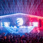 Crowd enjoying a nighttime concert with vibrant laser lights and colorful stage effects under a large canopy.