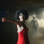 Woman in a red dress reaching out hand in a dark room with mist and a shadowy figure in the background