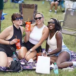 Three women wearing sunglasses enjoying drinks while sitting on a blanket at an outdoor event on grass.