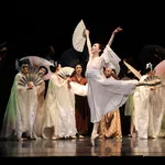 Ballet dancer in white costume performs with fans on stage with other dancers in traditional attire.