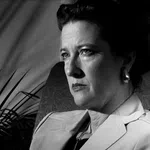 Black and white portrait of a serious woman sitting in a chair with shadows and a lamp in the background
