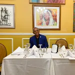 Man in blue suit sitting at white tablecloth-covered table set with wine glasses and silverware, framed art on yellow wall.