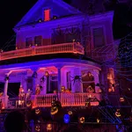 Large Victorian house decorated with glowing jack-o'-lanterns, skeletons, giant spiders, and web lights for Halloween night.