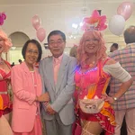 Two women in colorful circus costumes and pink wigs pose with a couple dressed in pastel pink and gray at a festive event.