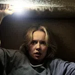 Woman trapped in a small dark space holding a bright flashlight above her head, looking fearful and tense.