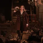 Man in historical coat carrying a wooden board with a dead animal in a dimly lit stone room with skeletons.