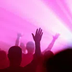 Silhouettes of people raising hands in a vibrant pink and purple light at a concert or event.