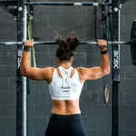 Woman lifting a heavy barbell overhead in a gym with a black brick wall background