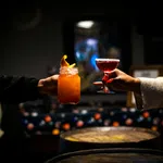 Two hands holding up colorful cocktails in dimly lit bar with wooden barrels in the foreground.