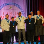 Group of chefs and officials posing with medals on stage at Gold Medal Plate culinary competition event.