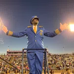 Band director in blue uniform conducting with baton at a stadium filled with spectators during evening.