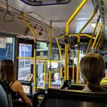 Passengers seated inside a city bus with yellow handrails and windows showing an urban street outside.