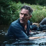 Two men cautiously wading through a river surrounded by dense greenery, holding onto a rope for support.