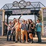Group of people posing and smiling under Sober Fest banner at outdoor event entrance on sunny day