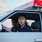 Female police officer and male colleague in a police car with red siren light, looking concerned through open window.