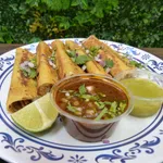 Plate of golden taquitos garnished with cilantro and onions, served with lime wedges and two dipping sauces.