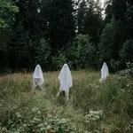 Three figures dressed in white ghost costumes standing in a grassy clearing surrounded by dense forest under overcast sky.