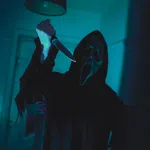 Person wearing a ghost face mask and dark robe holding a raised knife in a dimly lit room.