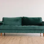 Modern emerald green velvet sofa with wooden legs on light wooden floor against plain white wall