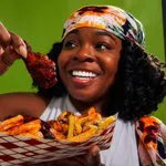 Smiling woman holding a saucy chicken wing and a tray of seasoned fries against a green background.