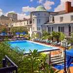 Outdoor pool area surrounded by lounge chairs, greenery, and historic white buildings under a blue sky.