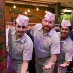 Four men wearing retro diner uniforms and hats posing with outstretched hands in a cozy restaurant setting.