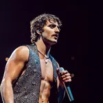 Muscular male singer with curly hair and sleeveless studded vest performing on stage with microphone