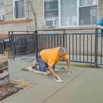 Construction worker smoothing fresh concrete on a sidewalk while another man watches at a residential building.