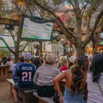 Outdoor sports bar with crowd watching a football game on multiple large screens under trees and string lights.