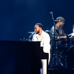 Singer playing piano and drummer performing on stage with dark blue background during concert