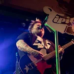Tattooed musician passionately playing electric guitar and singing into microphone on stage with vibrant lighting.