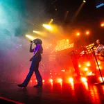 Live concert with a singer in a cowboy hat, guitarist, and vibrant colorful stage lighting and fog effects