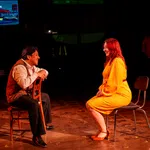 Man and woman in bright clothes sit facing each other on chairs in a dark theater setting with a screen in the background.