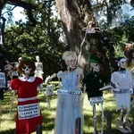 Halloween skeleton characters with humorous signs in a sunlit yard with trees and a house in the background.