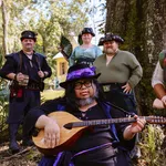 Group of six people in medieval and pirate costumes posing outdoors with a lute player in front under a large tree.
